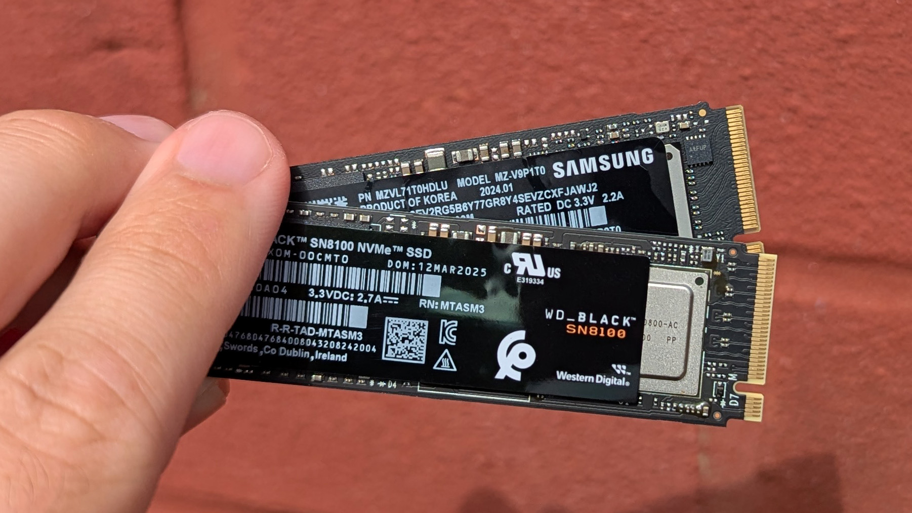 Close-up of a hand holding two NVMe SSD cards against a red brick background. The image highlights the labels and branding on the storage devices.