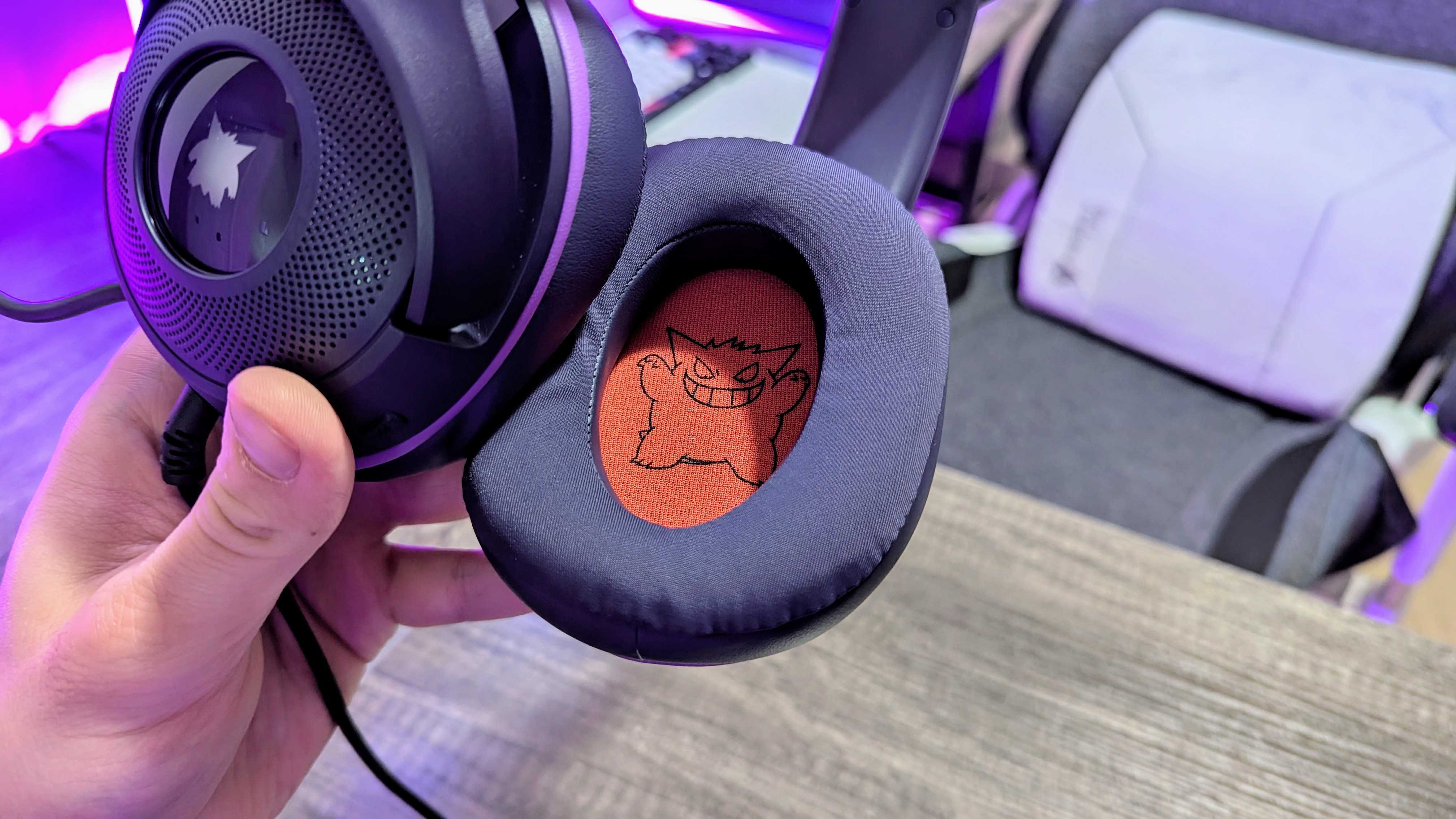 A hand holds black headphones with a mischievous cartoon character printed inside the ear cushion. A cozy chair and vibrant purple lighting are in the background.