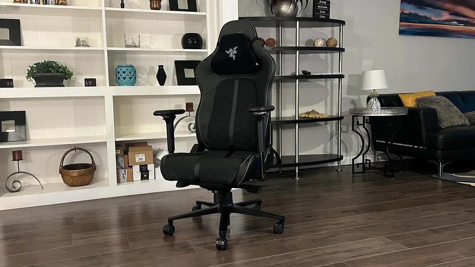 Ergonomic black gaming chair with armrests in a modern living room. Shelves with decor and a couch are in the background, creating a cozy vibe.