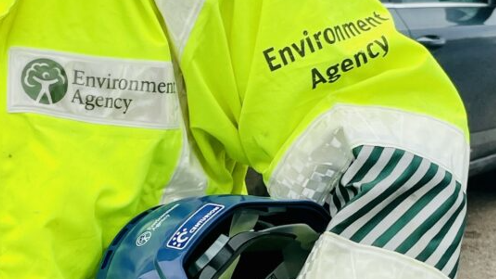 A close-up of a worker's arm in a high-visibility yellow jacket, featuring the Environment Agency logo and protective helmet.
