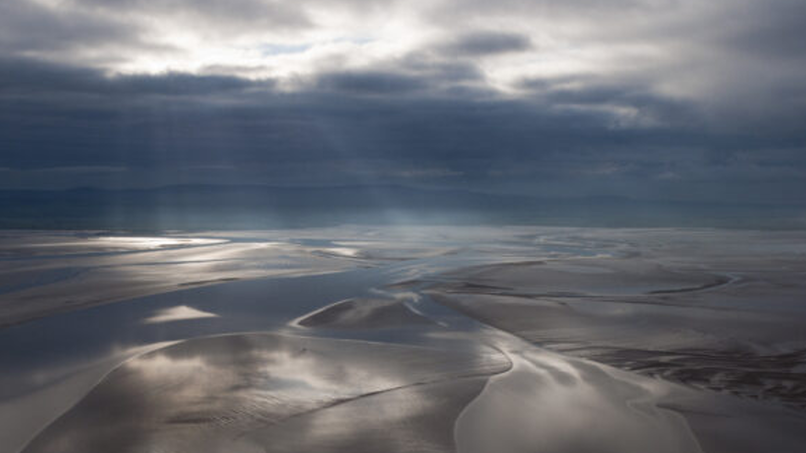A serene landscape of wet sand and shallow water reflecting stormy clouds, illuminated by soft rays of sunlight breaking through.