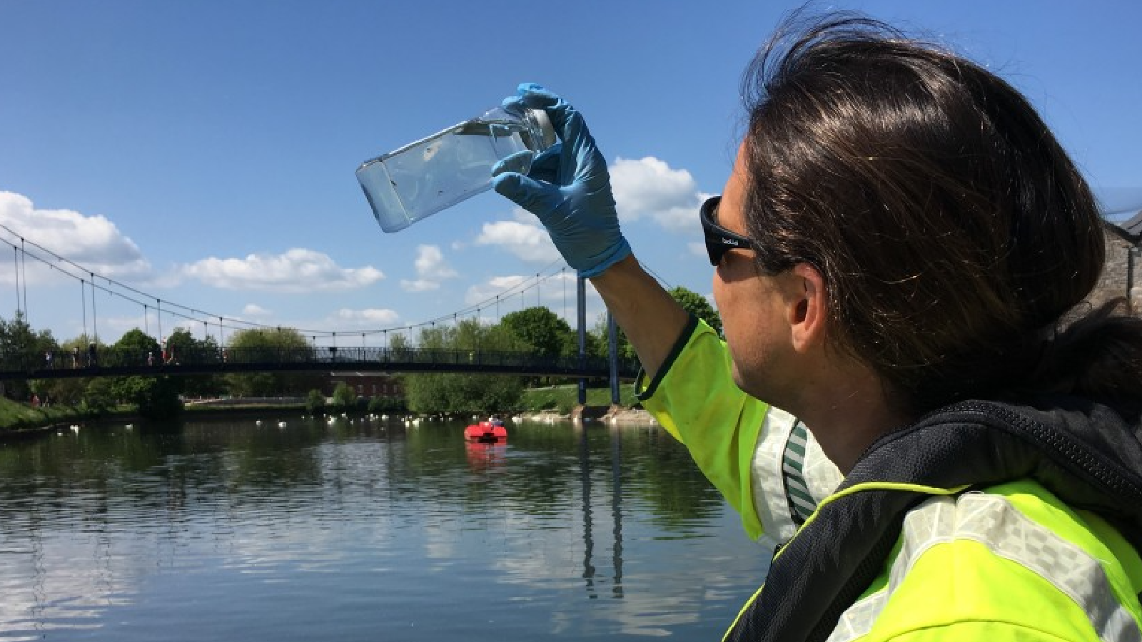 A person in a yellow jacket holds a plastic bottle over a river with a bridge in the background under a clear blue sky.