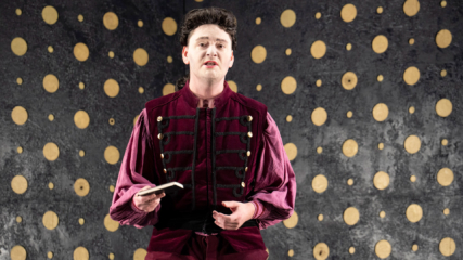 A curly-haired man in a red velvet jacket stands in front of an abstract background holding a book