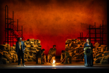 Male and Female chorus stand on either edge of the stage, looking at each other with concern. In the center of the stage, 3 performers dressed in military suits sit around a fire.
