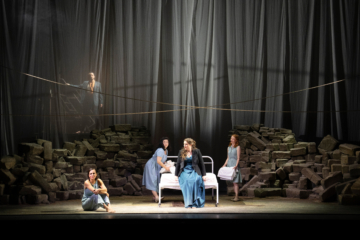 Female Chorus, Bianca and Lucia stand around a bed in the center of the stage and look over to Lucretia sitting on the floor. Male Chorus stands menacingly at the back of the stage behind a sheer curtain.