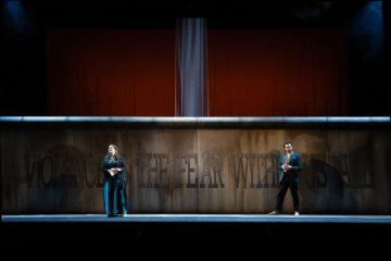 Male and Female Chorus stand in front of a tall concrete wall that says "violence is the fear within us all" in large letters.