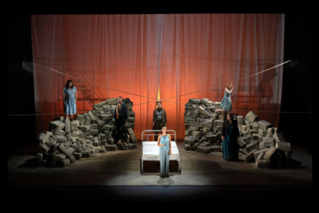 The cast of The Rape of Lucretia stands across the stage with a bed in the center and piles of rocks on either side.