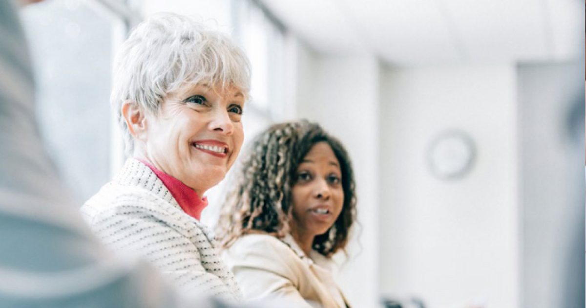 The image shows two women in a professional setting. The woman on the left has short gray hair, a light-colored jacket, and is smiling. The woman on the right has shoulder-length curly hair, a light-colored jacket, and appears engaged in conversation....