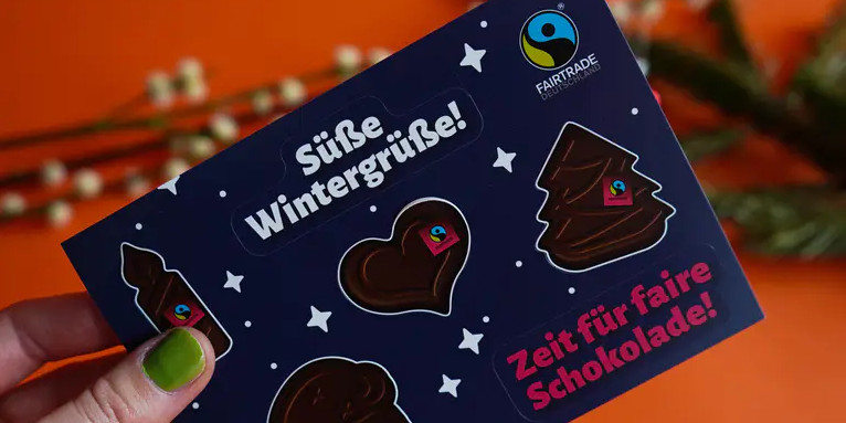dunkel blaue Stickerpostkarte mit Schokomotiven, die das Fairtrade-Siegel tragen und Text: "Süße Wintergrüße!" und "Zeit für faire Schokolade!"