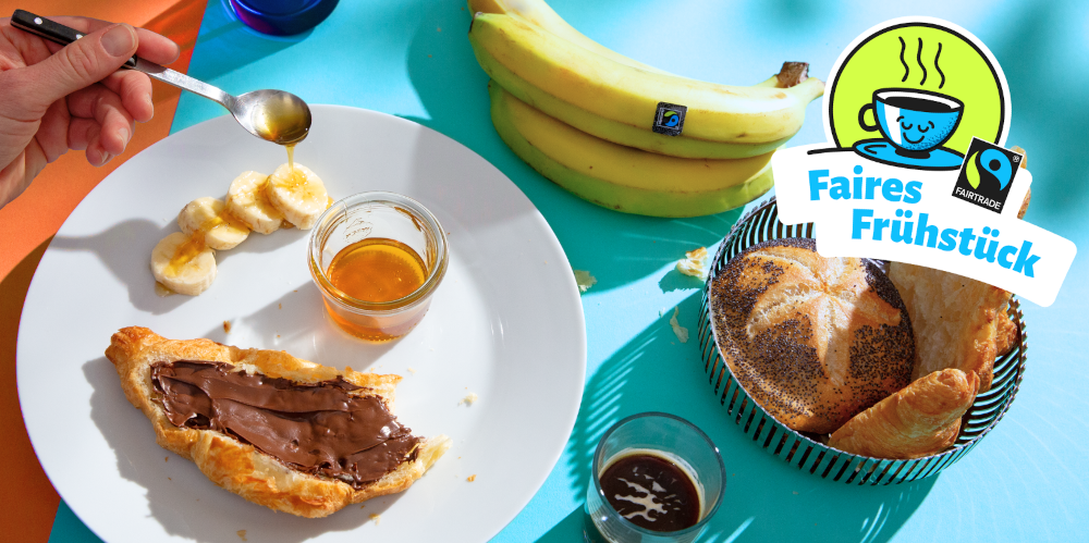 Bunter Tisch mit fairen Bananen, Brötchenkorb, Kaffeeglas, Honig und Croissant mit Schokoaufstrich
