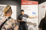 Ein Mann spricht vor einer Ausstellungswand mit Informationen zu einer Gruppe und gestikuliert dabei.