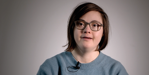 Eine junge Frau mit Brille und braunem, kinnlangen Haar sitzt vor neutralem Hintergrund und trägt ein Ansteckmikrofon