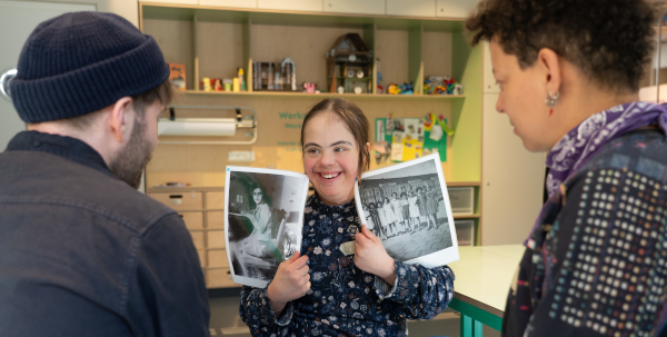 Frau zeigt zwei historische Fotos von Anne Frank; zwei weitere Personen von hinten blicken zu ihr