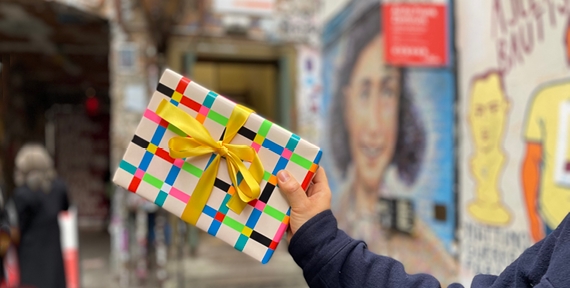 Eine Hand hält ein bunt verpacktes Geschenk mit gelber Schleife in die Höhe, während im Hintergrund das Wandportrait von Anne Frank unscharf zu sehen ist.