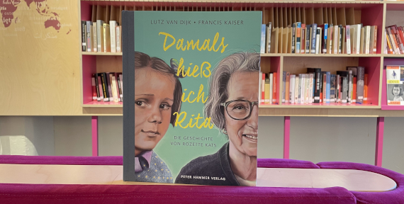 Buchcover mit dem Titel „Damals hieß ich Rita“ und den Porträts eines Mädchens und einer älteren Frau vor einer Bücherwand