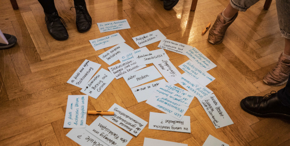 In der Mitte eines Stuhlkreises, auf einem Holfußboden, sind mehrere Moderationskarten mit Stichpunkten ausgebreitet.