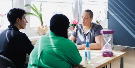 Drei Personen sitzen an einem Tisch im Gespräch, auf dem Tisch steht eine Sanduhr.