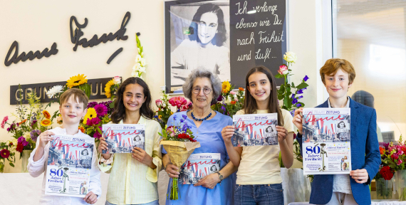 Eine Frau und vier Kinder stehen lächelnd nebeneinander und halten Zeitschriften hoch, während hinter ihnen ein großes Porträt von Anne Frank zu sehen ist.