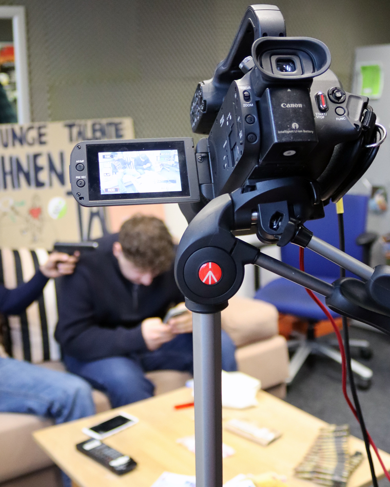 Das Bild ist auf eine Kamera im Vordergrund fokussiert, während eine Aufnahmesituation im unscharfen Hintergrund stattfindet. Die Filmkamera ist auf einem Stativ befestigt und auf einen dahinter liegenden Tisch und ein Sofa gerichtet. Darauf sitzen zwei nur unscharf erkennbare Personen, eine tippt am Handy und die andere hält einen Gegenstand in der Hand, der einer Schusswaffe ähnelt. Die Kamera fängt das Geschehen ein.