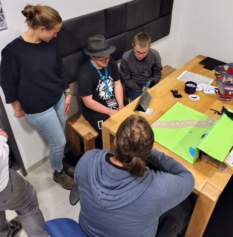 Das Foto zeigt zwei junge Erwachsene mit Downsyndrom beim Erstellen eines Stop-Motion-Films mit einer Action-Figur. Sie sitzen vor einer gebastelten Kulisse an einem Holztisch. Medienpädagogin Anna-Clara Pentz steht neben ihnen und erklärt ihnen, worauf es zu achten gilt.