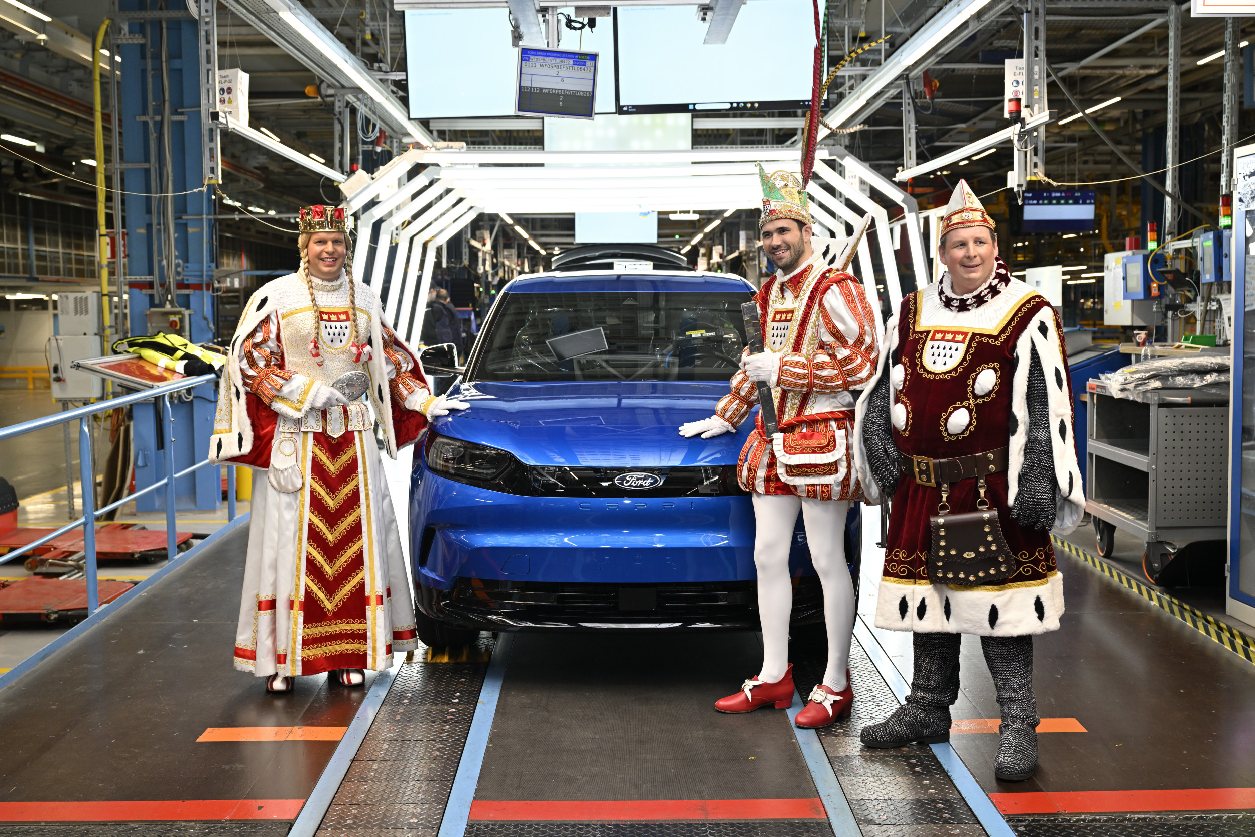 Drei für den Karneval verkleidete Männer stehen vor einem blauen Elektrofahrzeug der Marke Ford in einer Produktionshalle