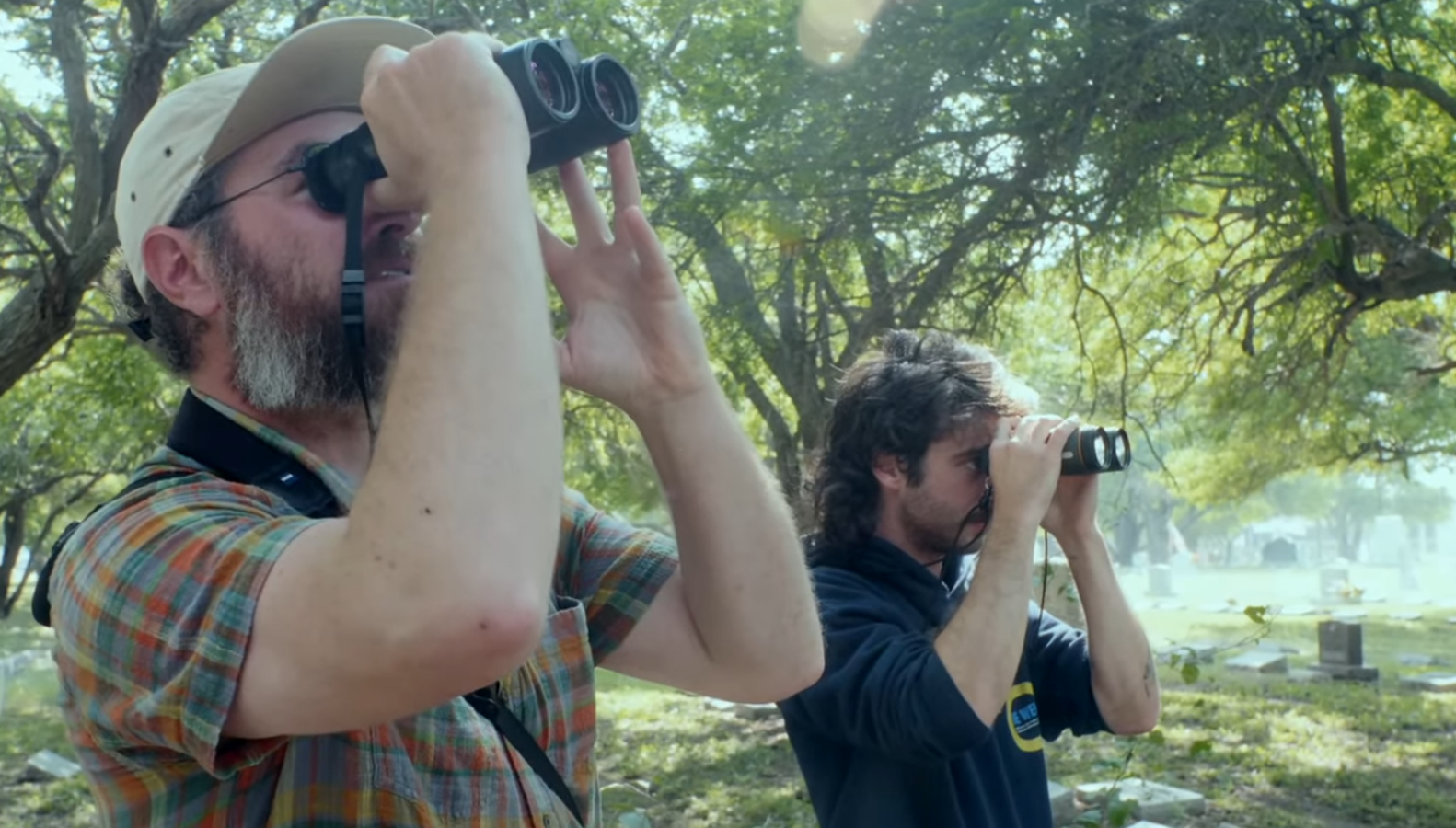 Zwei Männer blicken durch ihre Ferngläser. Im Hintergrund sieht man Grabsteine und einzelne Bäume.