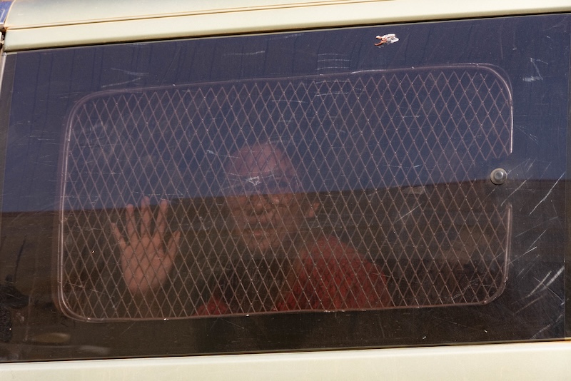 Eine Person sitzt im Inneren eines Fahrzeugs hinter einem stark verdunkelten Fenster mit gitterartiger Struktur und legt die Hand flach gegen die Scheibe, sodass nur ihr leicht verschwommenes Gesicht und die Hand durch das dunkle Glas erkennbar sind.