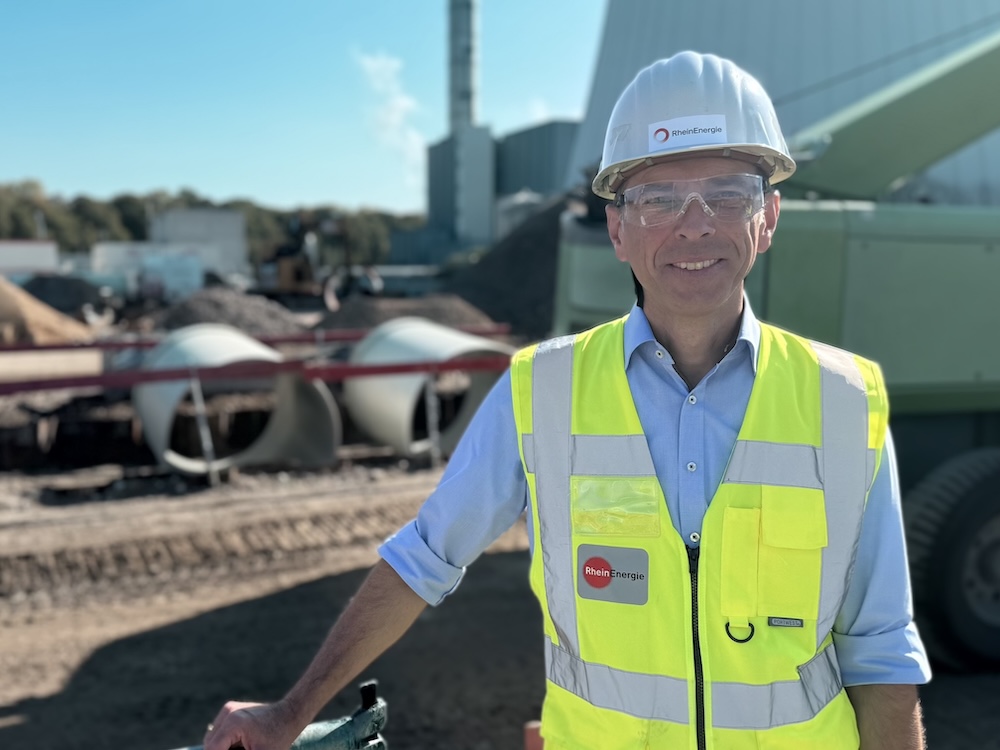 Porträt von freundlich schauendem Mann mit Schutzbrille und gelber Warnweste auf einer Baustelle; im Hintergrund liegen große Rohre und technische Anlagen.