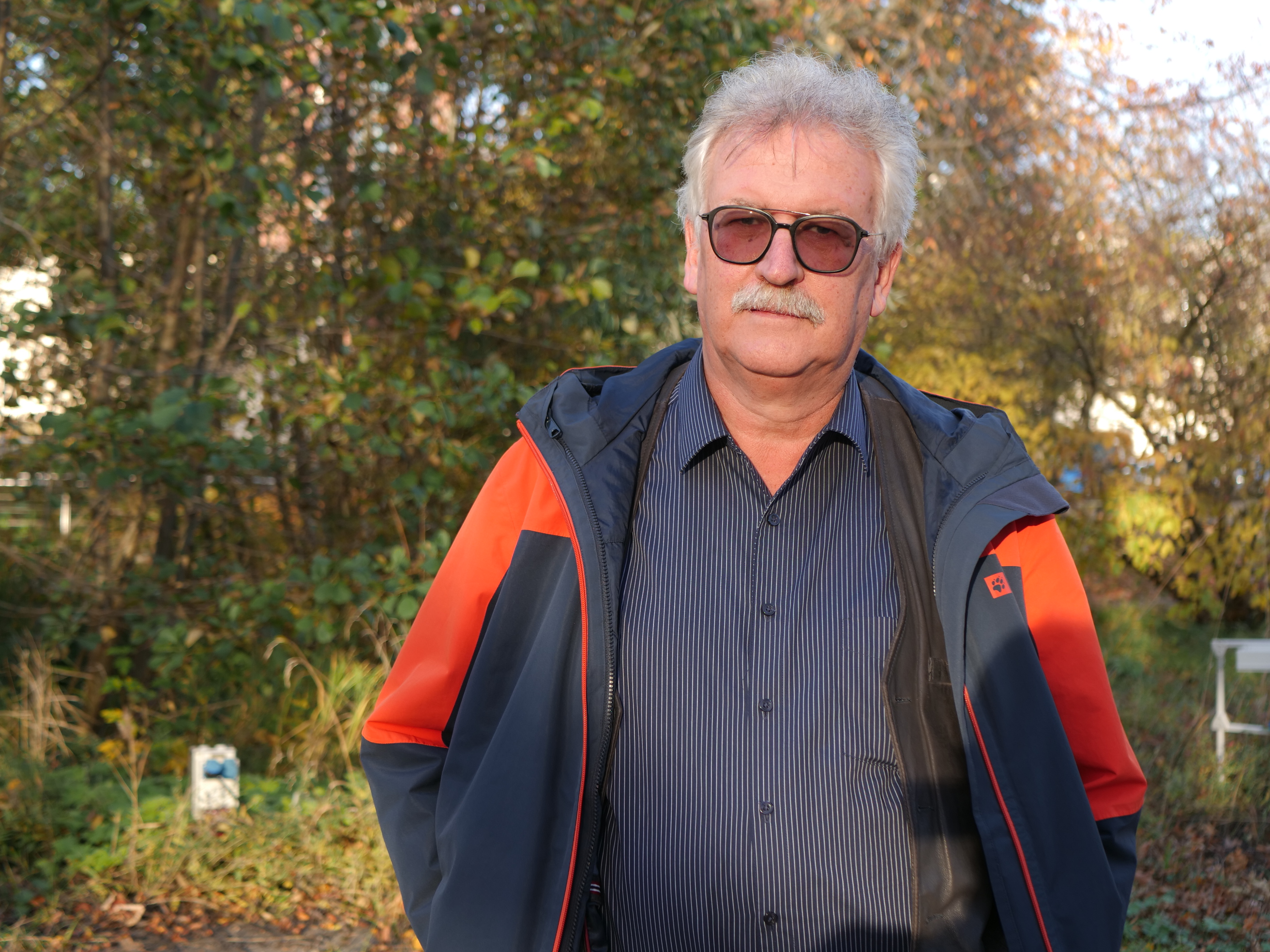 Ein Mann mit grauen Haaren, Schnurrbart und einer getönten Sonnenbrille schaut in die Kamera.