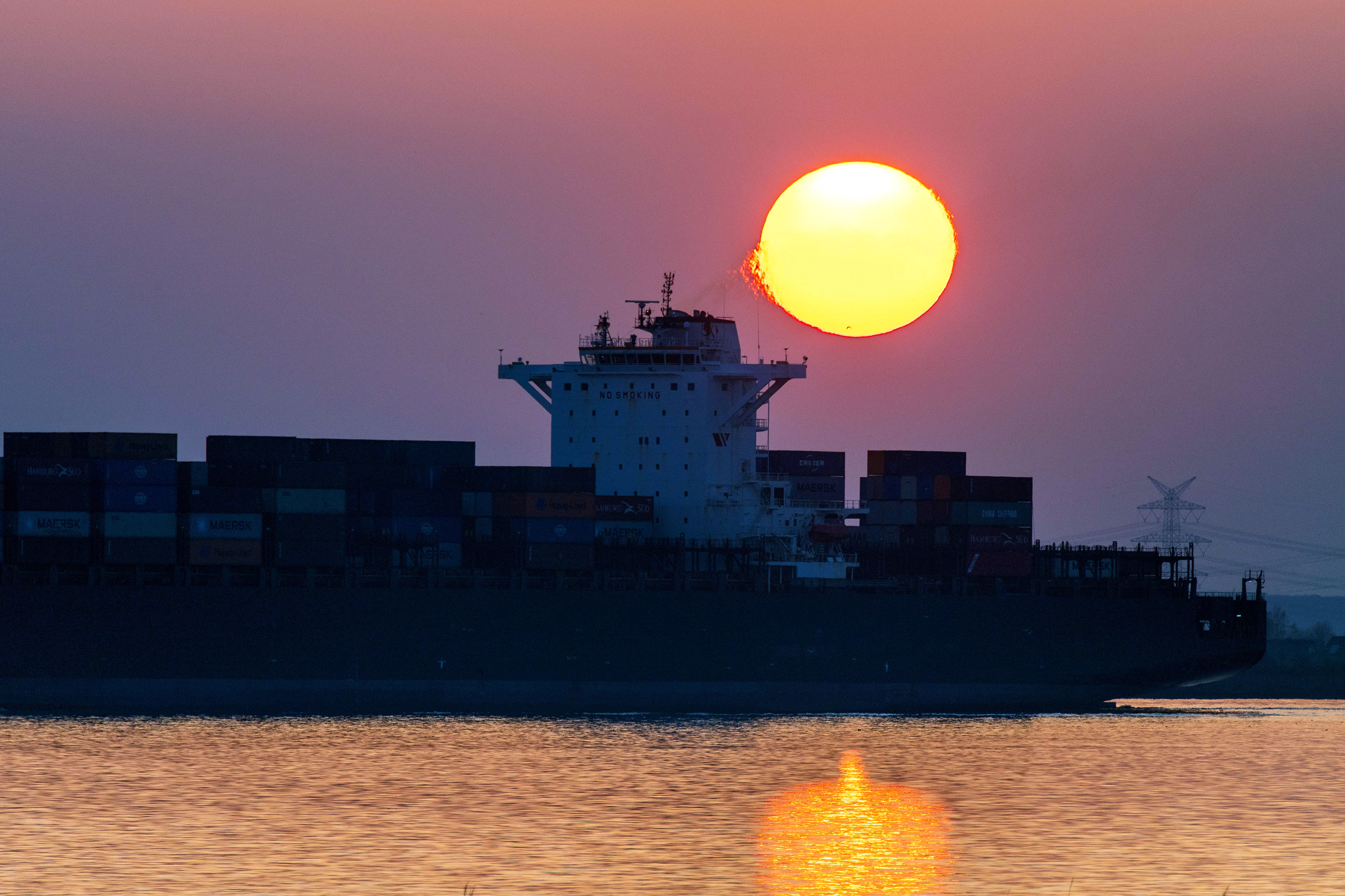 Ein Containerschiff auf der Elbe vor einer verschwommen rot leuchtenden Sonne.