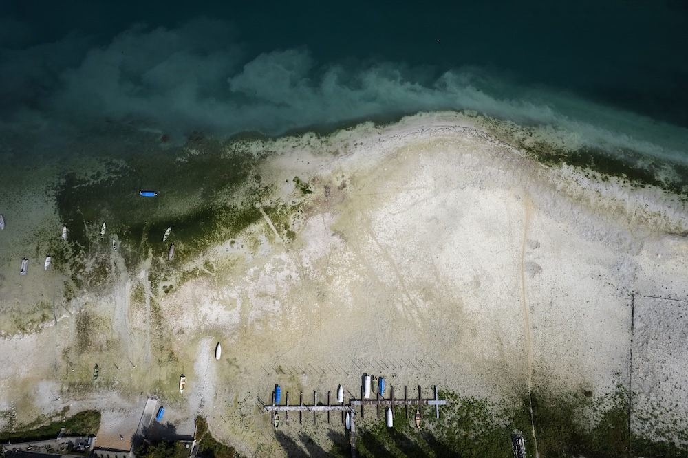 Drohnen-Luftaufnahme eines flachen Uferbereichs mit Booten, Stegen und türkisfarbenem Wasser. Einige Boote liegen in Reih und Glied auf dem Trockenen.