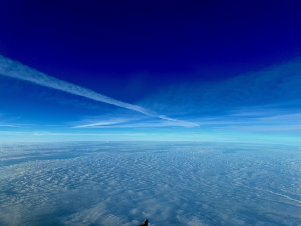 Luftaufnahme eines klaren blauen Himmels mit mehreren kreuzenden weißen Kondensstreifen von Flugzeugen; darunter eine geschlossene Wolkendecke aus lockeren, wellenförmigen Wolken in Hellblau; kontrastreiche Perspektive aus Flugzeug-Cockpit