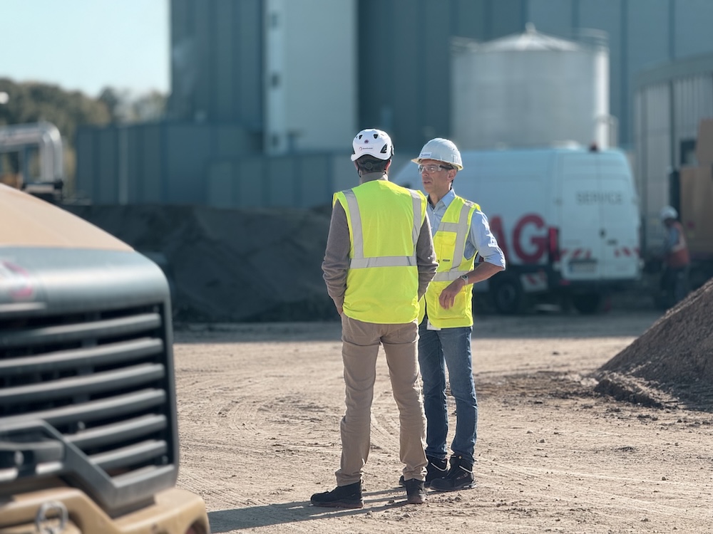 Zwei Mitarbeitende mit Schutzhelmen und Warnwesten sprechen auf einer Baustelle vor Industrieanlagen und Baustoffhaufen.