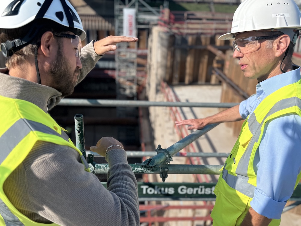 Zwei Mitarbeitende mit Schutzhelmen und Warnwesten sprechen auf einer Baustelle vor einem Bauloch mit Gerüsten.