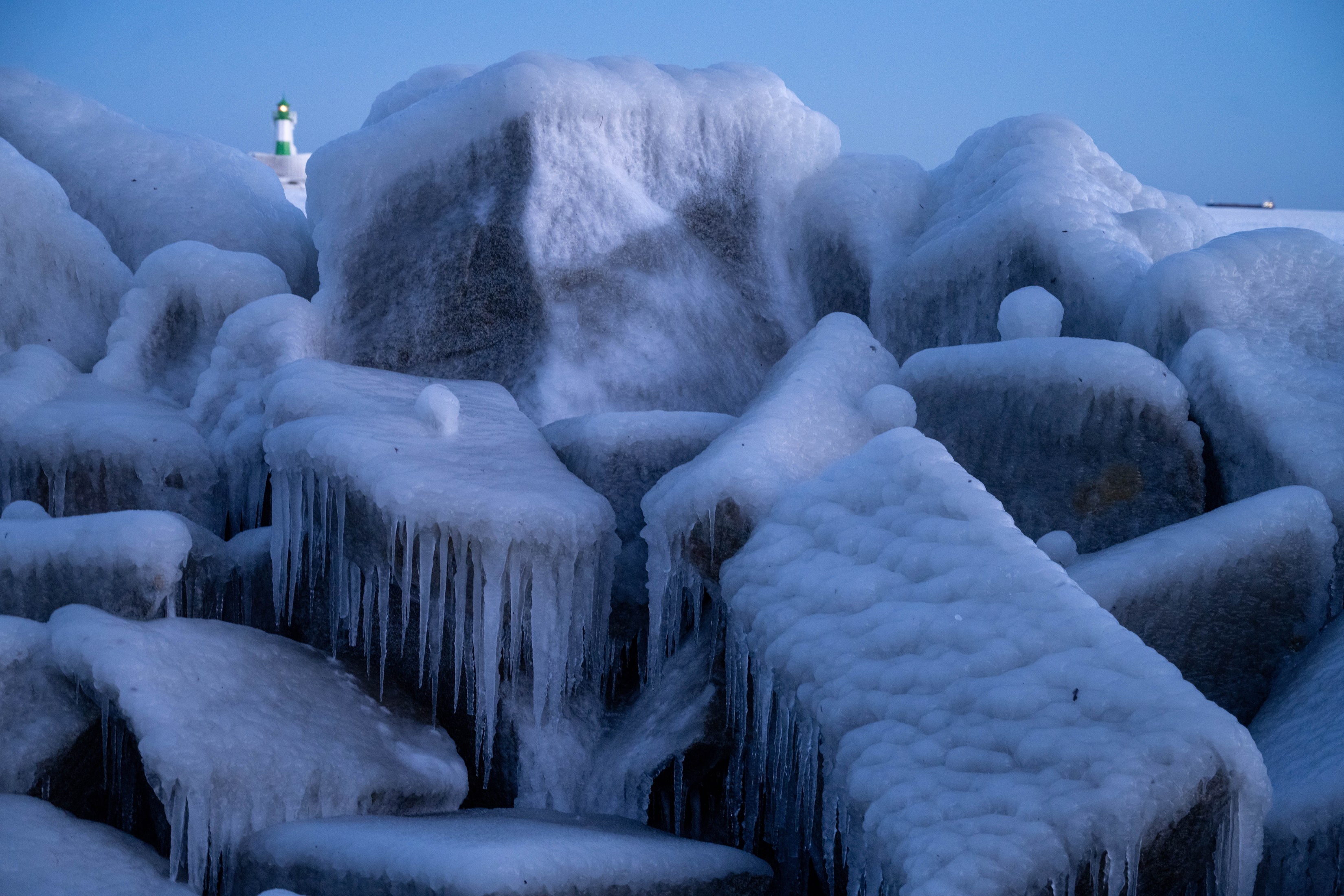 Eine dicke Eisschicht mit Eiszapfen auf Steinen, im Hintergrund der Leuchtturm von Sassnitz.