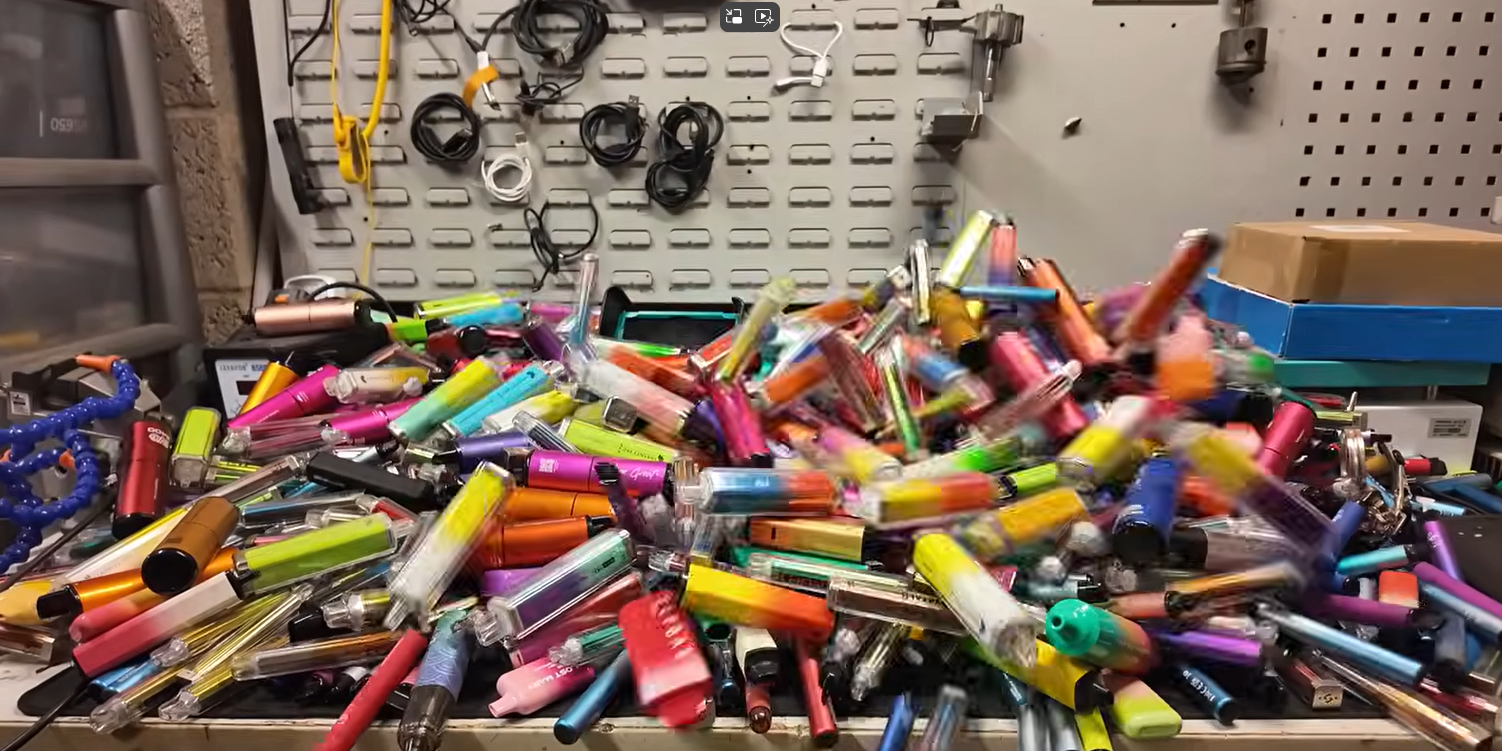 Viele bunte ausrangiertes Einweg-E-Zigaretten liegen auf einem Haufen, im Hintergrund aufgewickelte Kabel an der Wand.