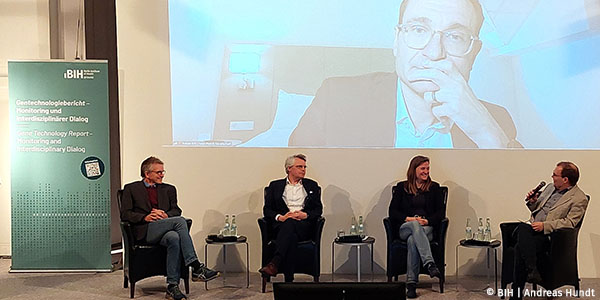 Panel der Veranstaltung: drei Männer und eine Frau sitzen auf dem Podium. Ein Mann hält ein Mikrofon und ist der Frau zugewandt. Die anderen beiden Männer schauen zu den beiden. Hinter den Personen ist auf einer Leinwand ein weiterer Gesprächspartner zugeschaltet. Links von den Sitzenden ist ein  Rollup mit Infos.