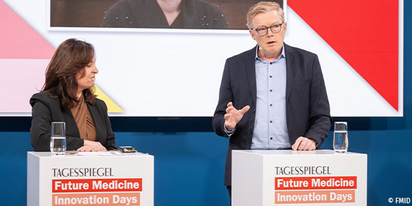 Professor Christopher Baum auf dem Podium der Future Medicine Innovation Days vor einem Rednerpult, auf dem ein Glas Wasser steht. Links von ihm steht  Ayse  Asar, MdB, ebefalls am Rednerpult, auf dem auch ein Glas Wassr steht.