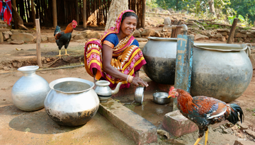 Eine junge indische Frau spült an einer Wasserstelle. NEben hier stehen Hühner und leere Wasserkanister