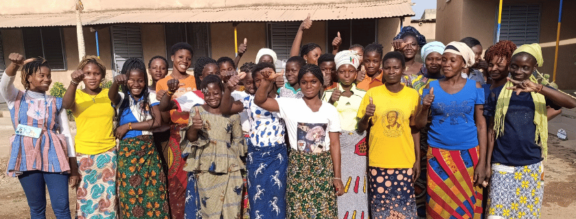 Eine Gruppe von ca 15 Frauen aus Burkina Faso vor einem Haus mit kraftvollem Blick und erhobenen Armen