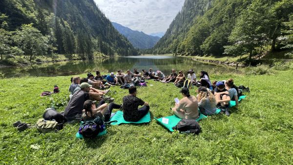 25 Seminarteilnehmende sitzen vor einer sommerlichen Bergkulisse am Bergsee.