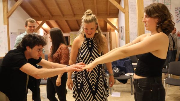 Ein junger Mann und eine junge Frau halten die Fingerspitzen aneinander. Im Hintergrund steht eine weitere Frau und schaut interessiert zu.