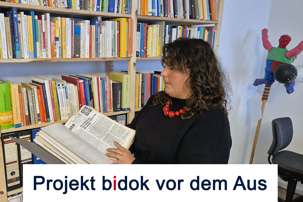 Projekt-Leitung Andrea Urthaler steht vor einer Bücherwand und hält eine Mappe mit Zeitschriften in ihren Händen. Sie hat dunkelbraunes lockiges Haar und trägt eine Kette mit großen roten Holzperlen.