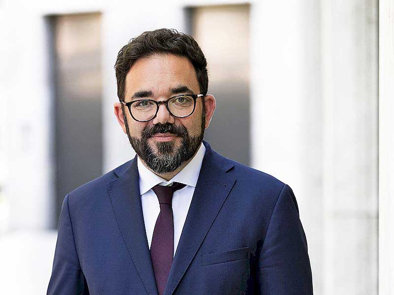 Porträtfoto: Ein Mann mit schwarzen kurzen Haaren, Bart und Brille blickt freundlich in die Kamera. Er trägt ein weißes Hemd, dazu eine auberginefarbene Krawatte und ein blaues Sakko.