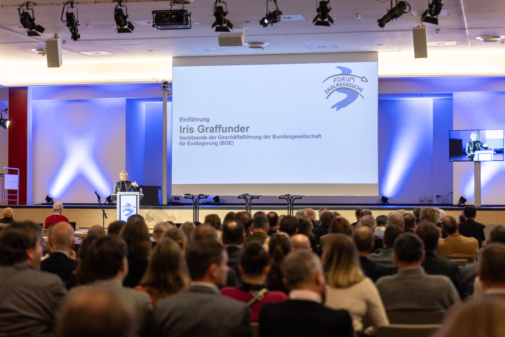 Auf mehreren hintereinander aufgestellten Stuhlreihen sitzen Personen, die von hinten zu sehen sind und auf eine Bühne blicken. Dort steht an einem Redner*innenpult eine Person. 