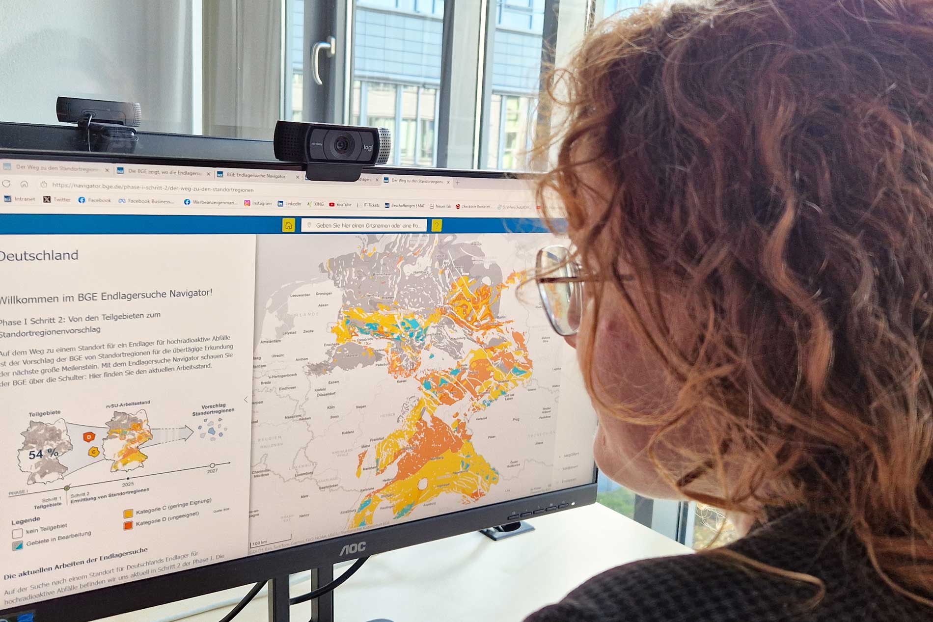 Vor einem Bildschirm, der eine Deutschlandkarte mit farbig markierten Flächen zeigt, sitzt eine Frau mit Brille und Locken. Sie ist schräg von hinten zu sehen. 