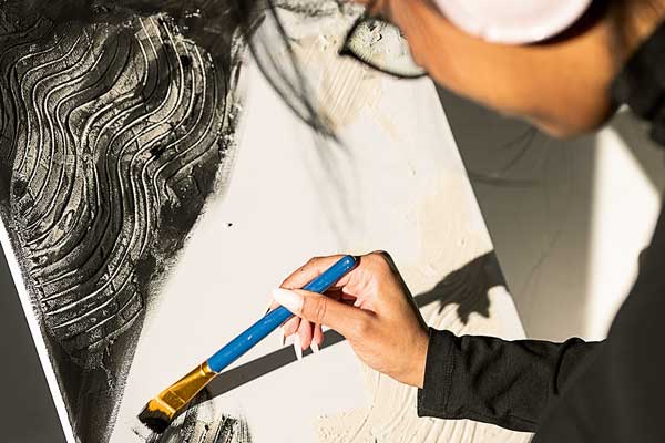 Das Bild zeigt eine Frau im Anschnitt, die mit einem Pinsel auf einer Leinwand malt.