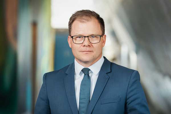 Porträtfoto: Ein Mann mit kurzen Haaren und Brille blickt frontal in die Kamera. Er trägt ein weißes Hemd, dazu eine blaue Krawatte und ein blaues Sakko.