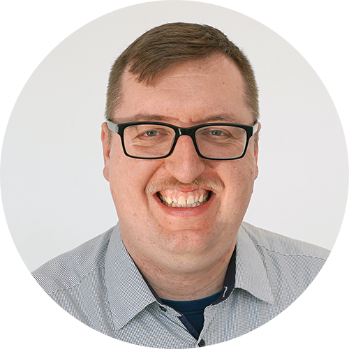 Ein Mann mit kariertem Hemd, schwarzer Brille und blondem Oberlippenbart lächelt in die Kamera.