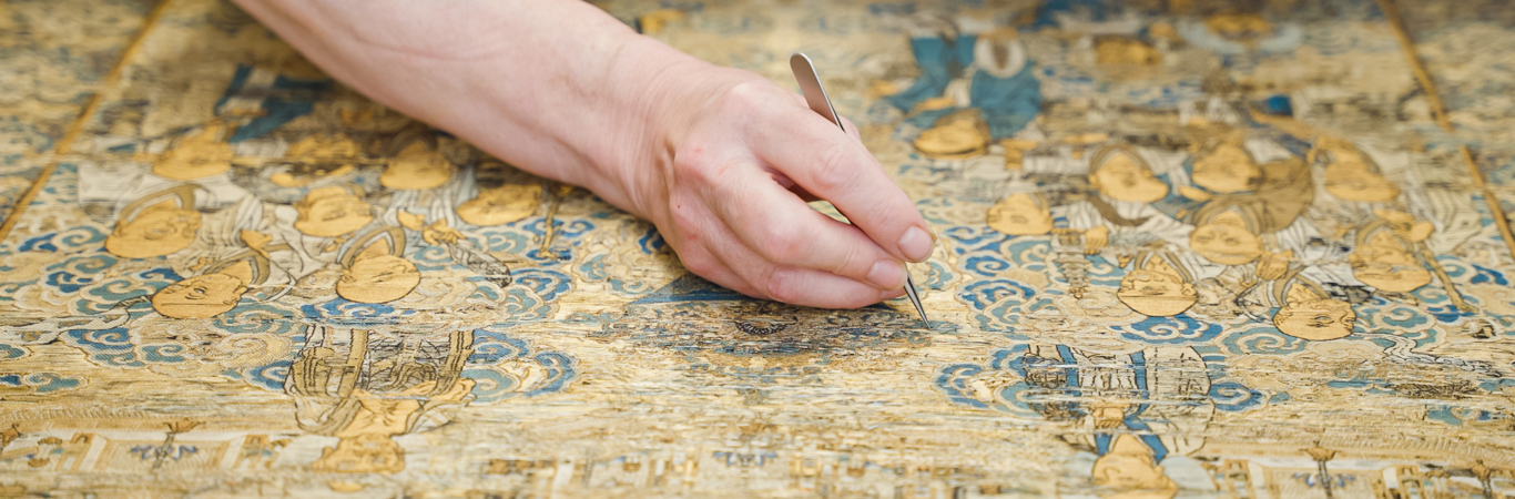 Hand holding a tweezers, leaning over an intricate gold and blue tapestry